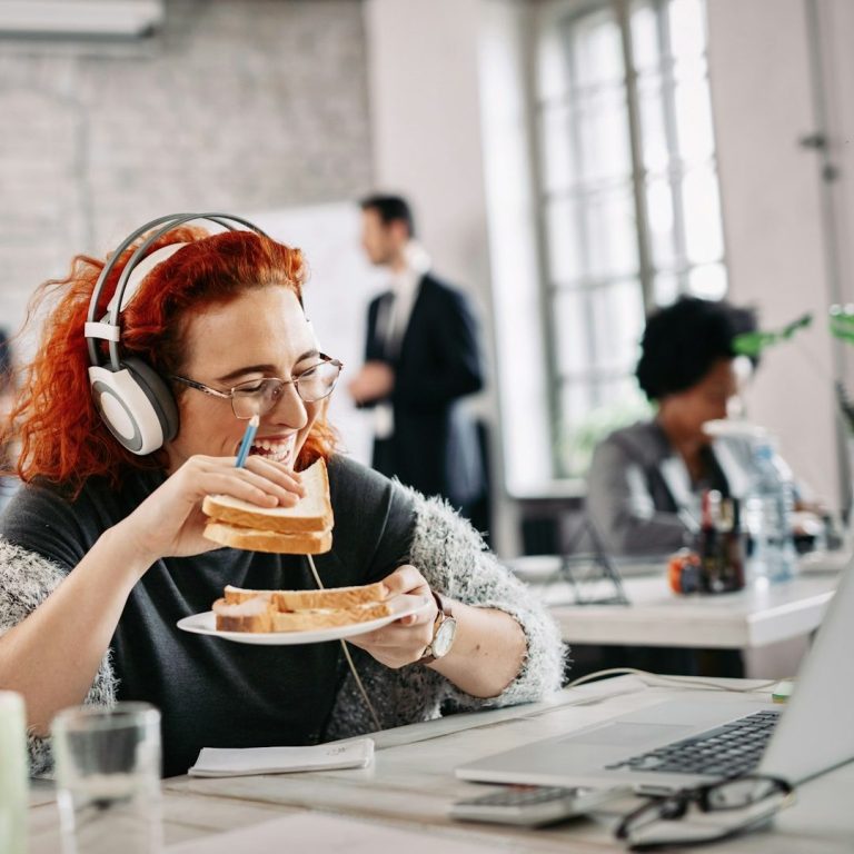 Frau mit roten Haaren und Headset auf dem Kopf, im Büro, isst ein Sandwich